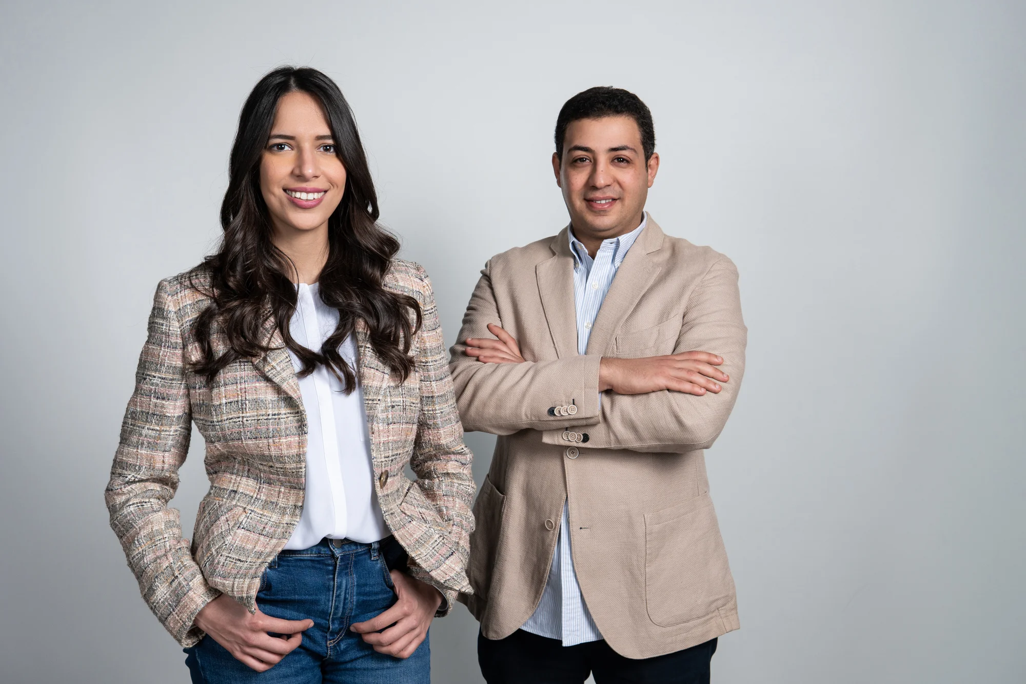 A woman in a tan plaid blazer and long brown hair stands next to a man wearing a beige blazer and white shirt in front of a grey background.