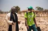 Two men walk across a dusty field, with trees in the background. One of the men is wearing a green t-shirt and green baseball cap, both with the OKO logo, along with an ID tag and a face mask. The other man is wearing a long white robe, a long black scarf around his neck and a gray kufi.