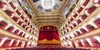 A picture of an opera house, taken from the middle seating area, showcasing the red and gold of stage curtains, large decorative ceiling chandelier, and the red and gold-framed four-storey boxes on both sides.