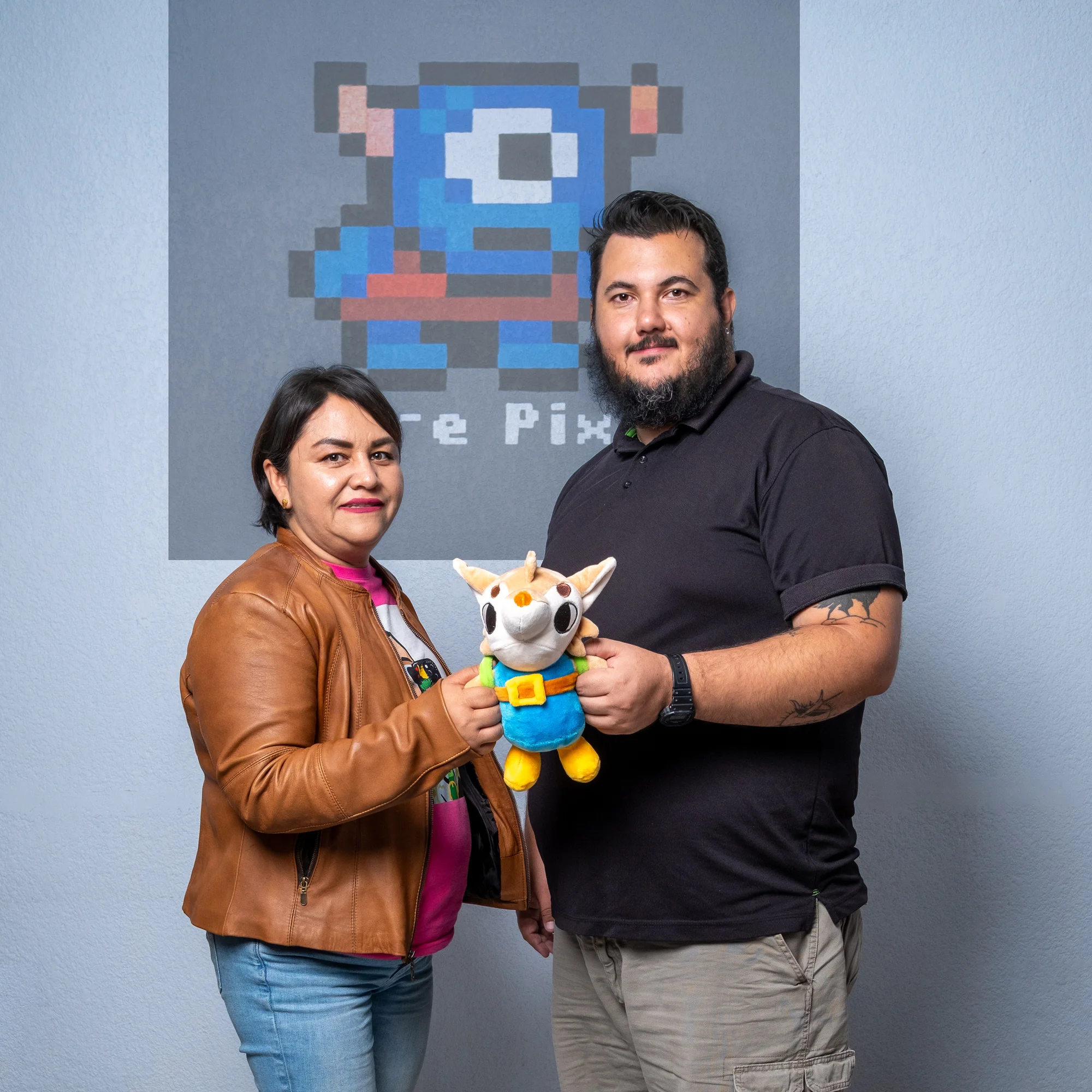 Two people stand in front of a sign with the Ogre Pixel logo  and hold a plush toy character.