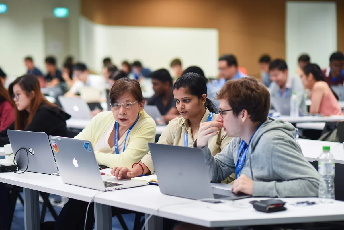 Three people sitting at a table with laptops, in a room full of people.