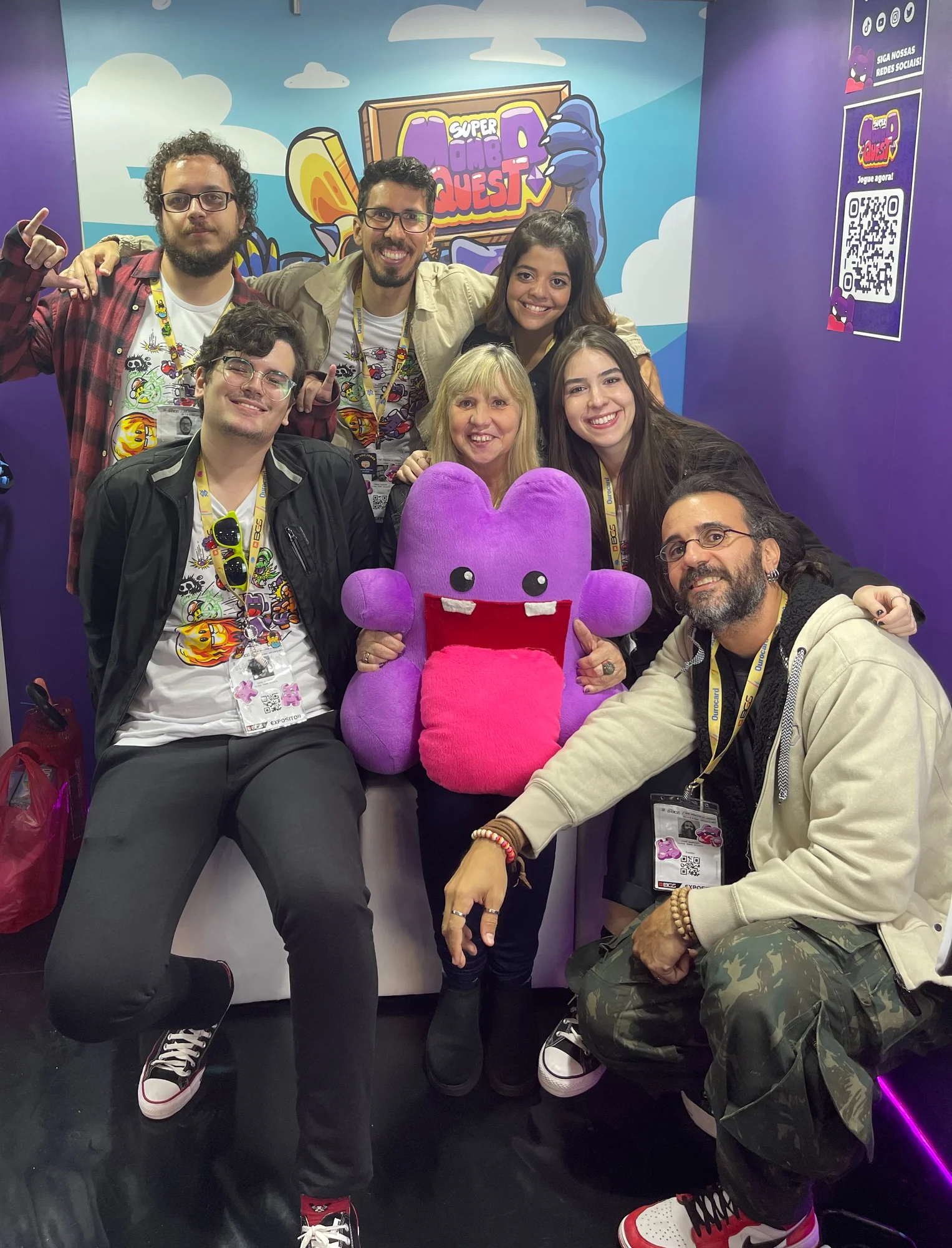 Seven people smile with a purple stuffed animal in a booth. In the background, a poster says “Super Mambo Quest.”