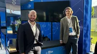 Two men in suits stand next to the company's booth at a conference.