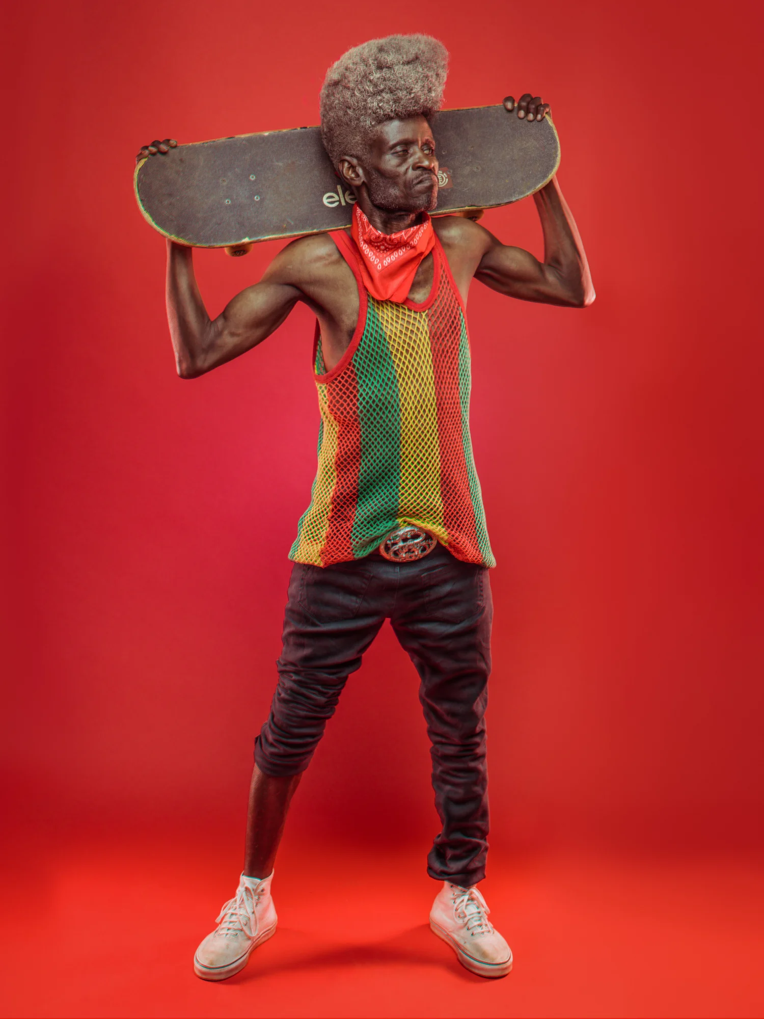 A man stands and holds a skateboard behind his head. He stands in front of a bright red background. He wears white shoes, black bants (rolled up on his right leg), and a green, yellow, and red-striped mesh top.