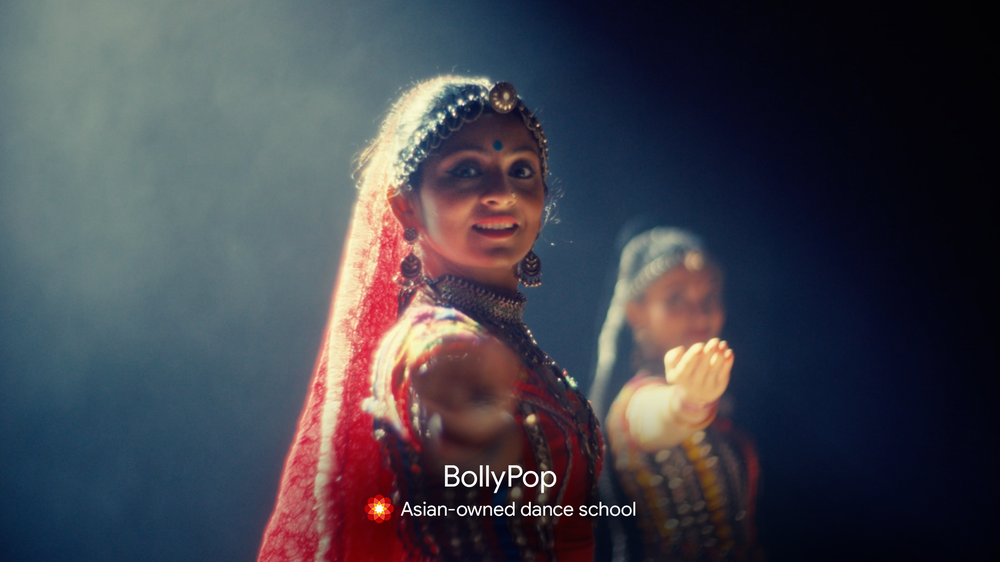 The owner of Asian-owned dance school, Bollypop, is standing, smiling towards the camera. She is extending her right arm and hand outward toward the camera in a dance pose. She is in red, traditional dress from Delhi, India. Her friend is behind her in th