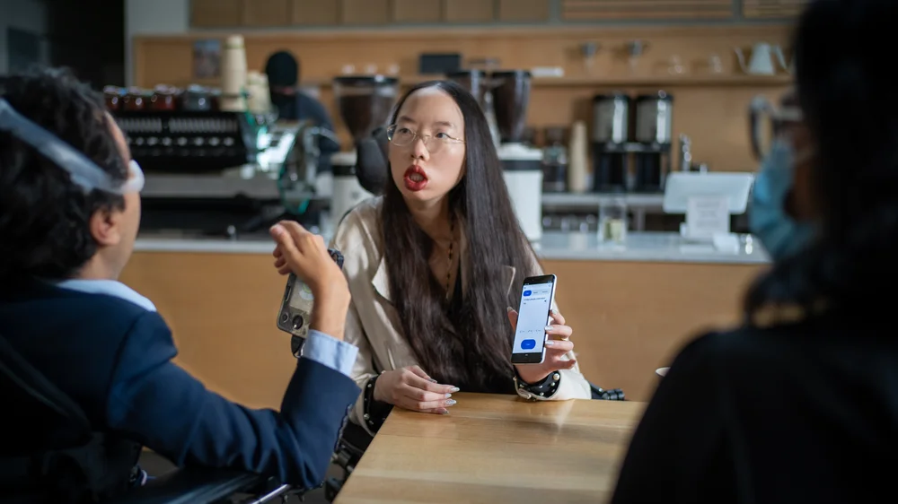 Image shows asian woman wearing glasses with long wavy dark hair and red lipstick. She is using a wheelchair and holding up a phone that displays the Project Relate app to a person across the table from her, who is wearing a suit and uses a wheelchair.