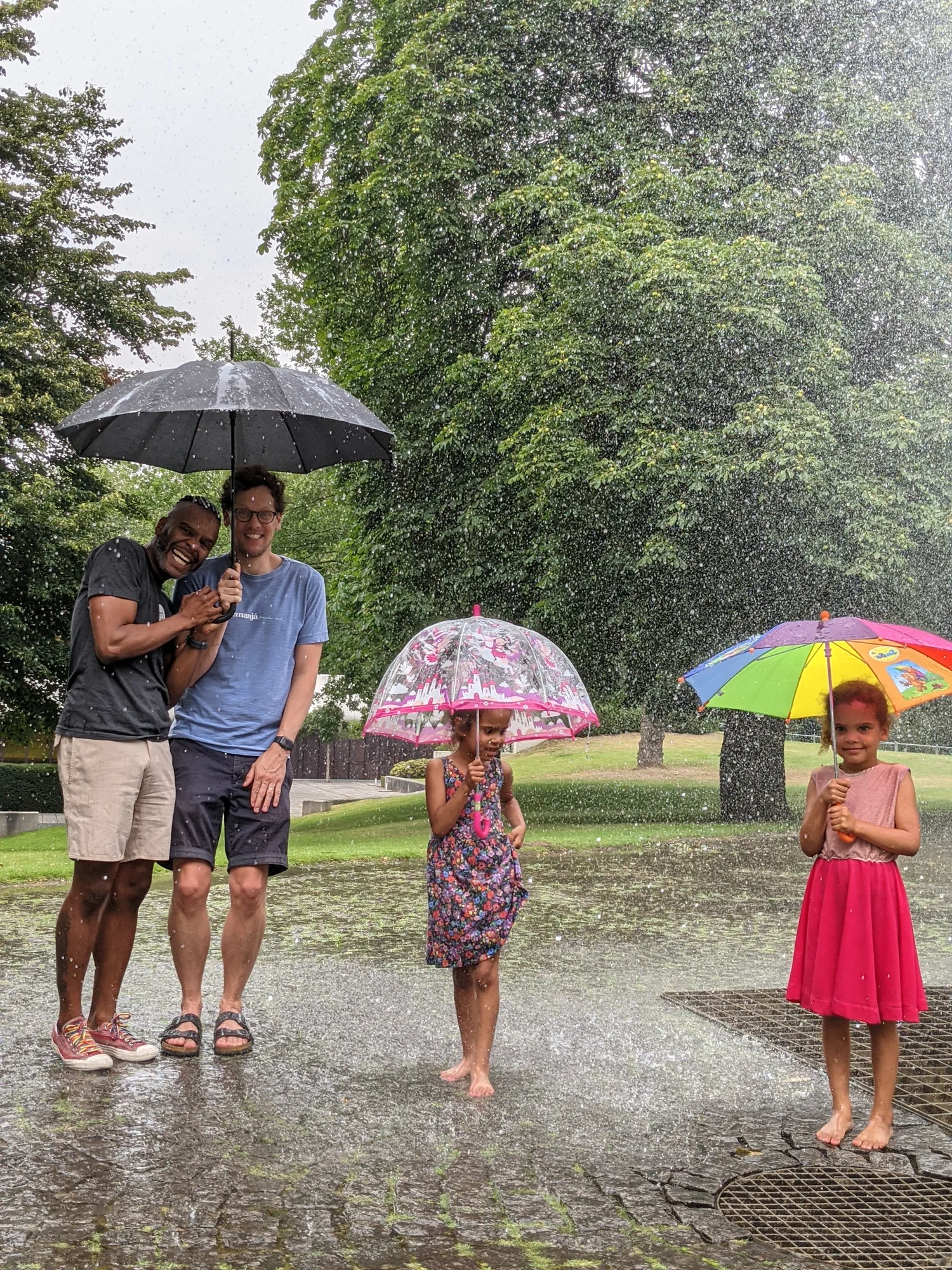 Daniel und sein Mann Ole stehen gemeinsam unter einem Regenschirm, es regnet stark. Sie tragen beide T-Shirt und kurze Hosen. Neben ihnen stehen ihre beiden Kinder, die jeweils auch einen Regenschirm halten. Alle schauen in die Kamera.