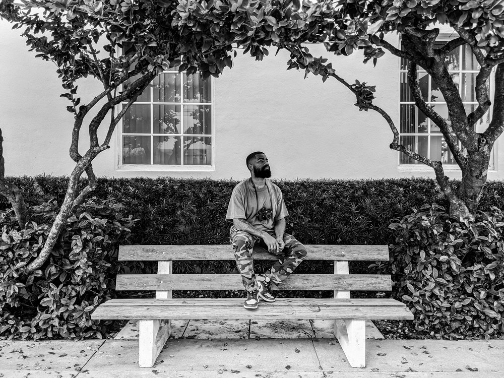 A black and white image showing a person sitting on a bench in between two trees. The person is sitting on top of the back of the bench looking up at the trees.