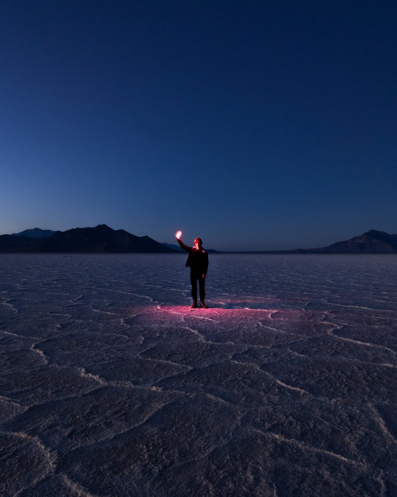 Image showing a person standing in a desert at dusk; the sky is dark blue. A small ping light is shining around him while he holds a lit up object in his hand.