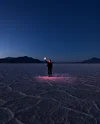 Image showing a person standing in a desert at dusk; the sky is dark blue. A small ping light is shining around him while he holds a lit up object in his hand.