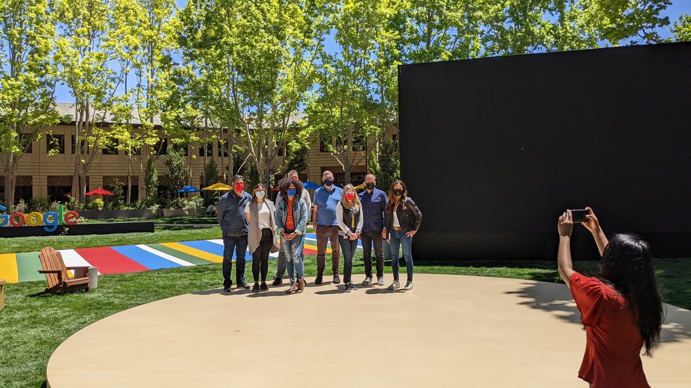 Photo showing a group of people wearing masks standing on a circular stage on a lawn. A person in the foreground is taking a photo of them.