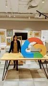 Chai, in a green shirt and black jacket, is standing behind a wooden table with a large Google Cloud logo sitting on it. Hanging on the wall in the background is Van Gogh’s self portrait, part of the exhibit she’s visiting.