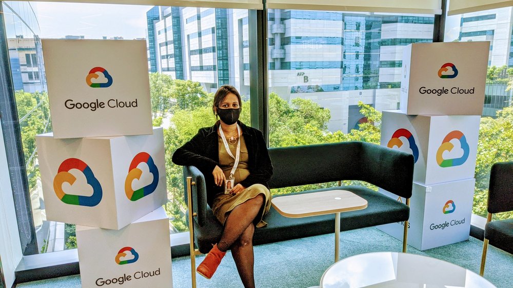 Chai, in a green shirt, black jacket, and black mask, is sitting on a couch in front of a window. On both sides of the couch are three stacked boxes that say “Google Cloud” and feature Cloud’s logo.