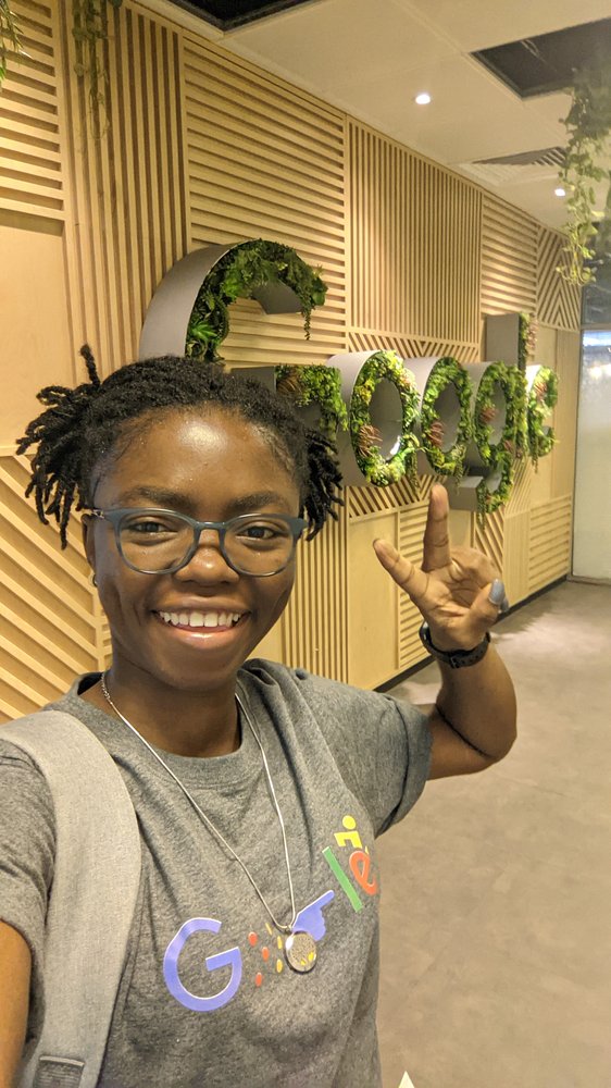 Oiza, wearing a Google t-shirt, smiles and holds up the two-finger “peace” sign in front of the Google logo.