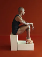 Photograph of a person sitting on a white box against a red backdrop. The person is wearing a one piece black clothing item and tall yellow high heels. They are facing away from the camera.