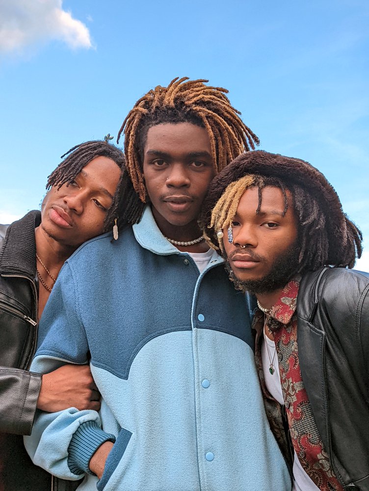 Photograph of three people standing together, hugging and leaning into one another. There is blue sky and white cloud in the background.