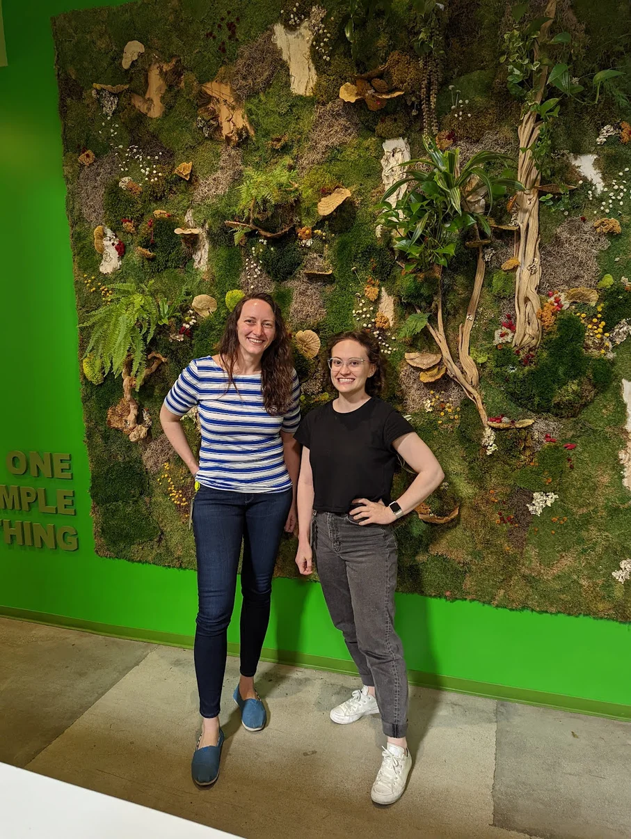 Two people stand side by side, looking into the camera and smiling. They’re standing in front of a green wall with a garden art installation on it.]