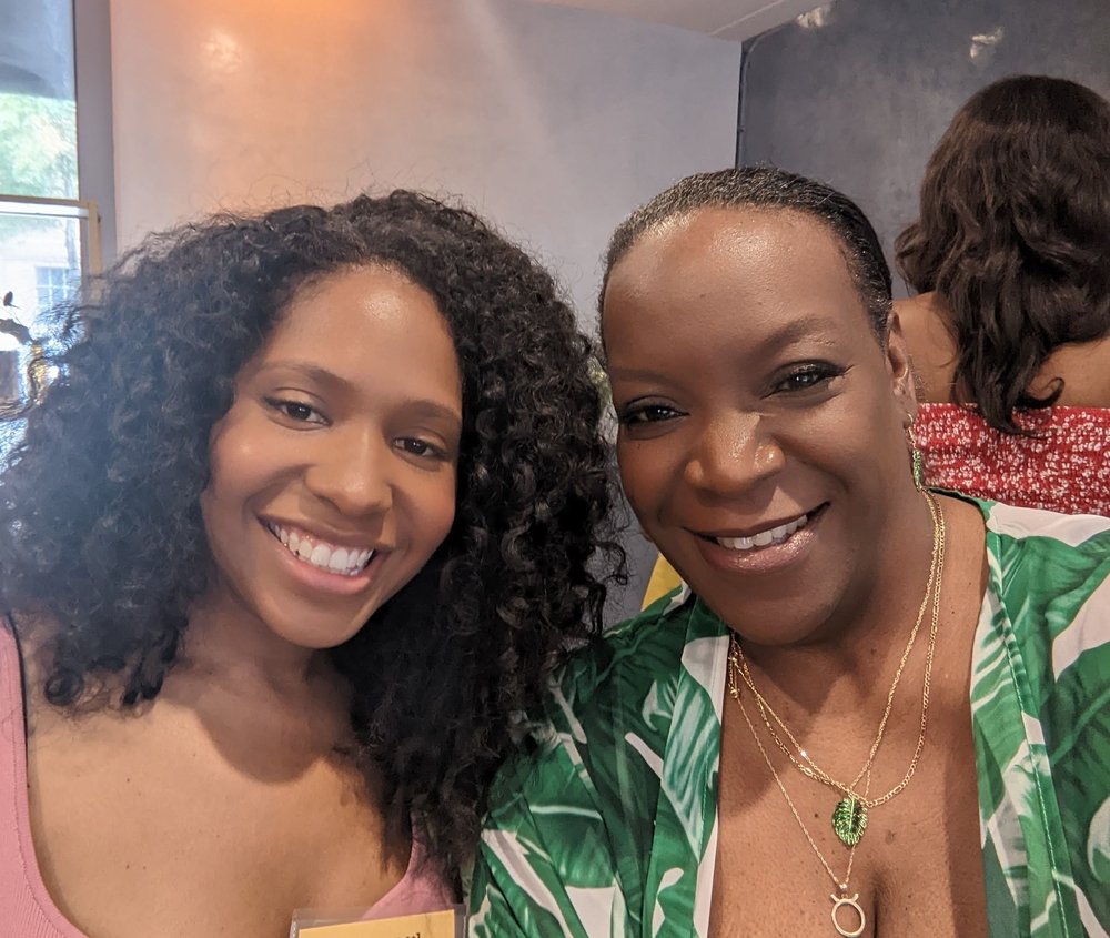 Two women smile at the camera — one wears a rose-colored top, another wears a green leaf print top and green jewel pendant necklace.