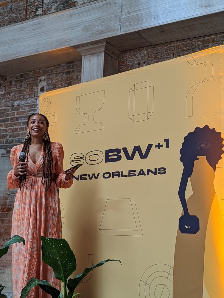 A woman in a long peach-colored patterned dress with a microphone stands in front of a large yellow sign that reads “SOBW +1 New Orleans.”