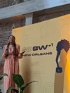 A woman in a long peach-colored patterned dress with a microphone stands in front of a large yellow sign that reads “SOBW +1 New Orleans.”