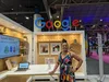 A woman in a multicolored dress stands in a booth under a Google sign, with her hand resting on a table.