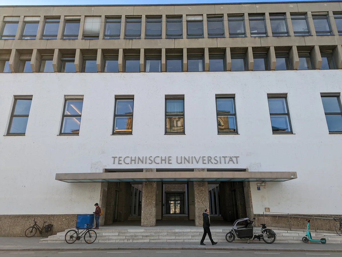 Unigebäude von außen, Treppe zum Eingang, darüber Fenster, 2 Passanten gehen vorbei