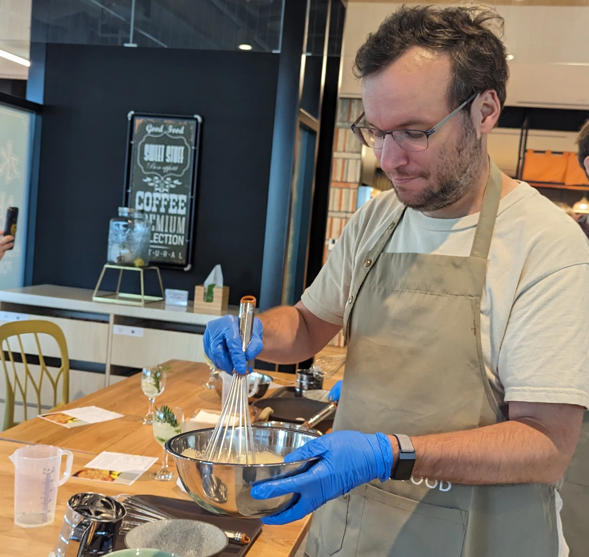 A person wearing glasses, a tan apron, and blue rubber gloves, holding a mixing bowl and whisk in a kitchen