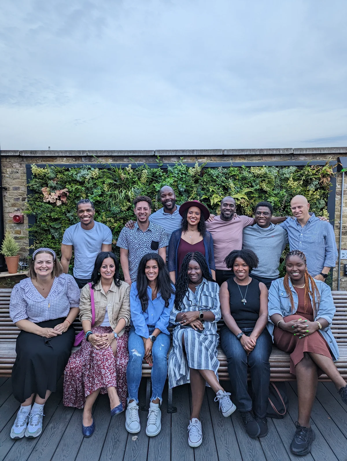 Thirteen individuals are pictured in two rows. The first row are seated and the second row are standing in front of a plant wall on a rooftop