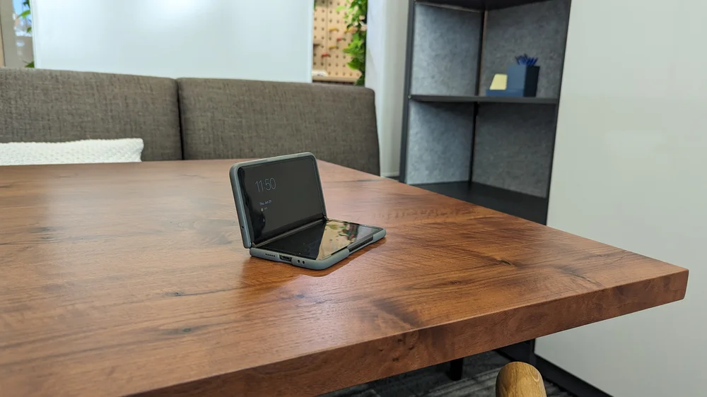 A photo of a Pixel Fold in tabletop mode, where it’s folded at the hinge so that one screen is upright and the other faces up at the ceiling so that upright screen is easily viewed. The Fold is sitting on a wood table in an office space.