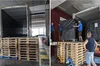 On the left, two stacks of pallets sit on the floor next to an empty refrigerated sea freight container. On the right, people move one of Google’s special book transportation carts from a refrigerated sea freight container over the two stacks of pallets.