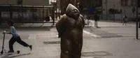 The photo shows a bronze statue of a small, chubby figure with a conical hat, standing on a city street. The statue appears to be one of the many "Wrocław dwarfs"  that are scattered throughout the city of Wrocław. In the background, there is a blurred image of a child riding a scooter, adding a sense of movement and daily life to the scene. The setting includes an urban street with a crosswalk and several people in the distance.