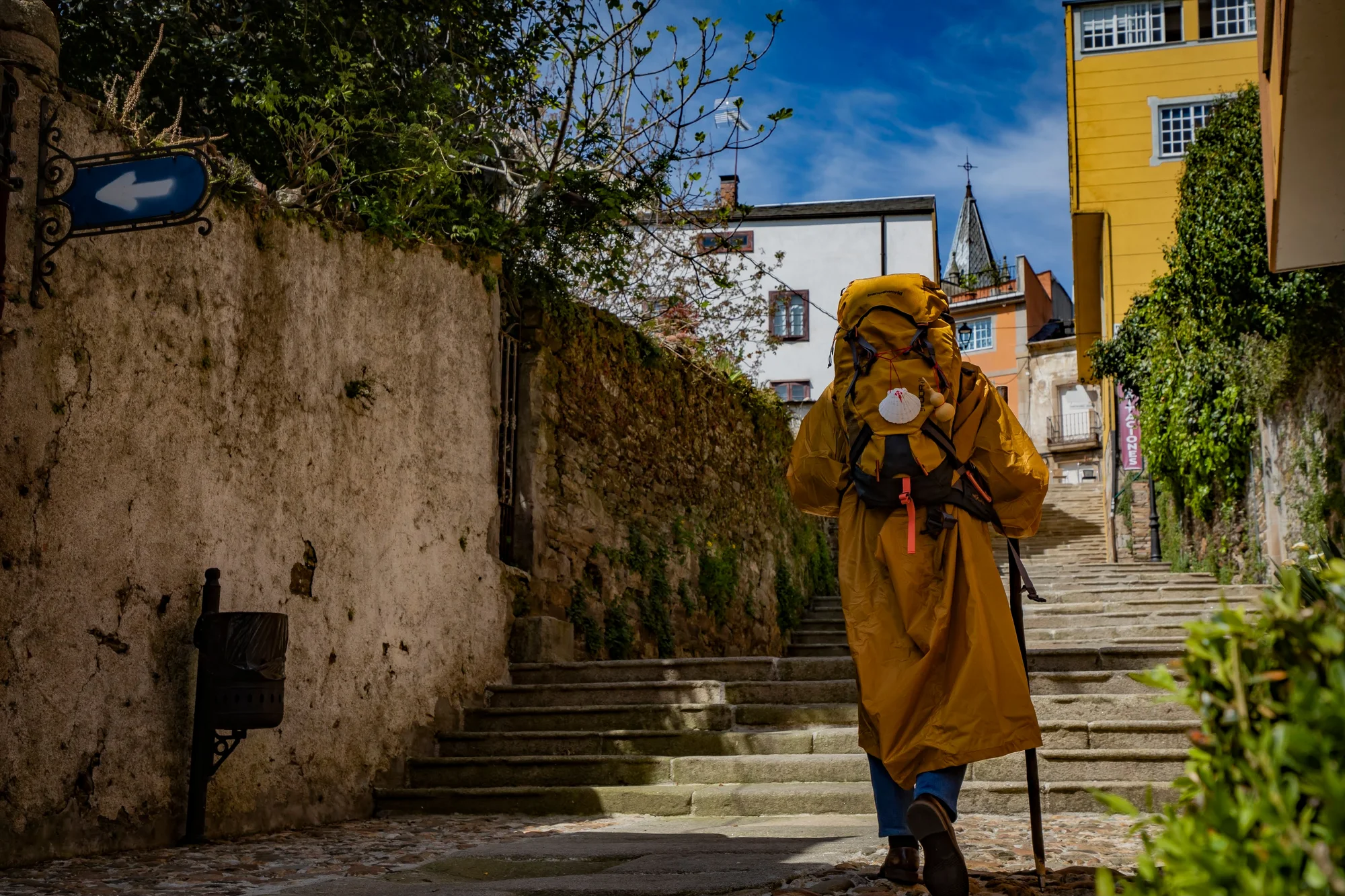 Photograph of a pilgrim with his back turned, wearing a yellow raincoat and a backpack. He is climbing some stairs on a narrow street.