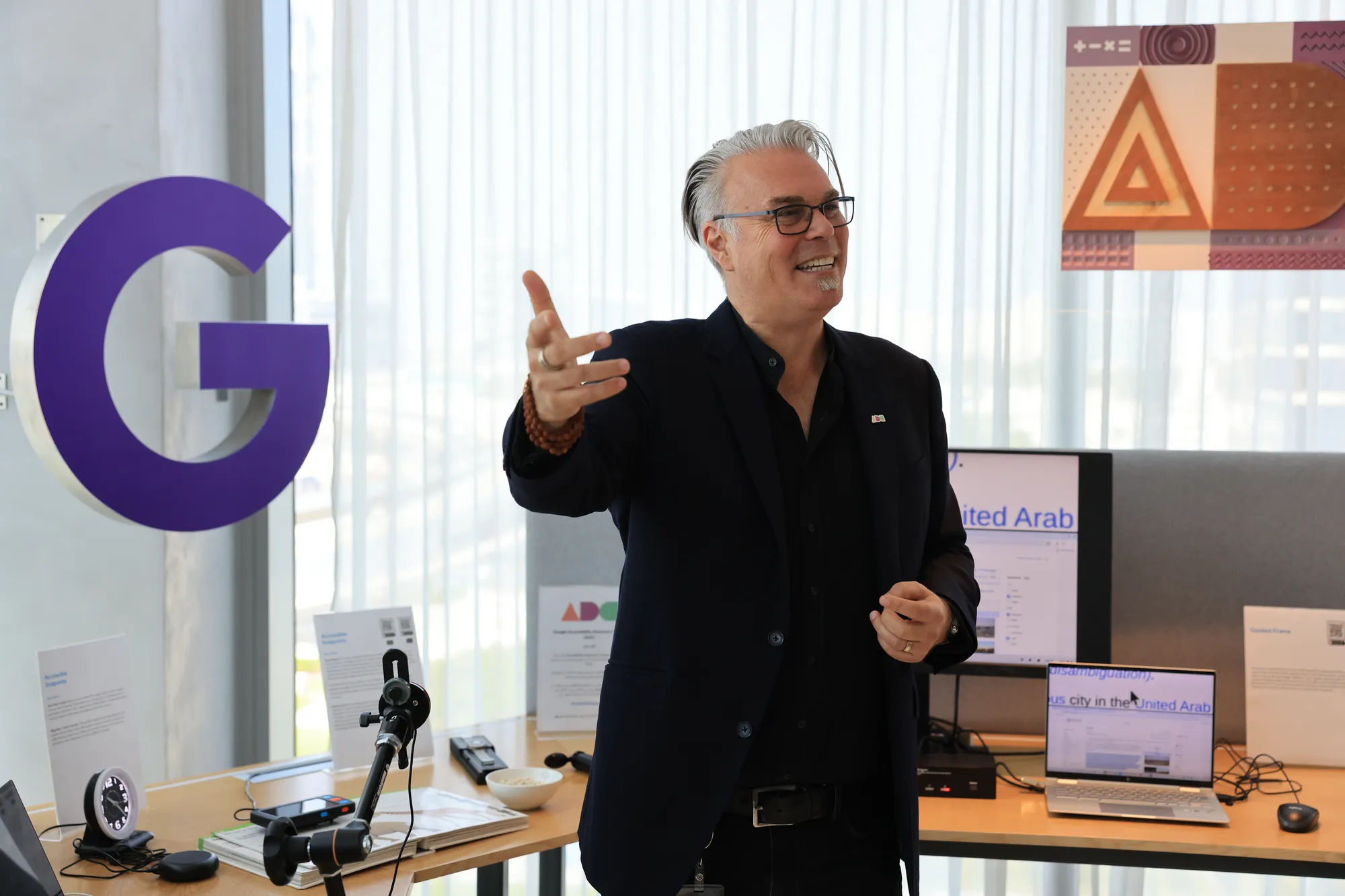 An image showing a man with blonde / greying hair gesturing to his right. He is wearing a black suit with a black shirt. Behind him is a wooden bench that features laptops and accessibility tools like a deaf alarm clock. There is also a large purple G hanging on the wall