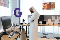 A man stands in front of a brown bench with a computer and accessibility switch buttons including a chin switch. On his right is another wooden bench with laptops and phones. Three posters on a gray fabric-covered wall. Each poster displays the Google Accessibility Discovery Center logo and Arabic text