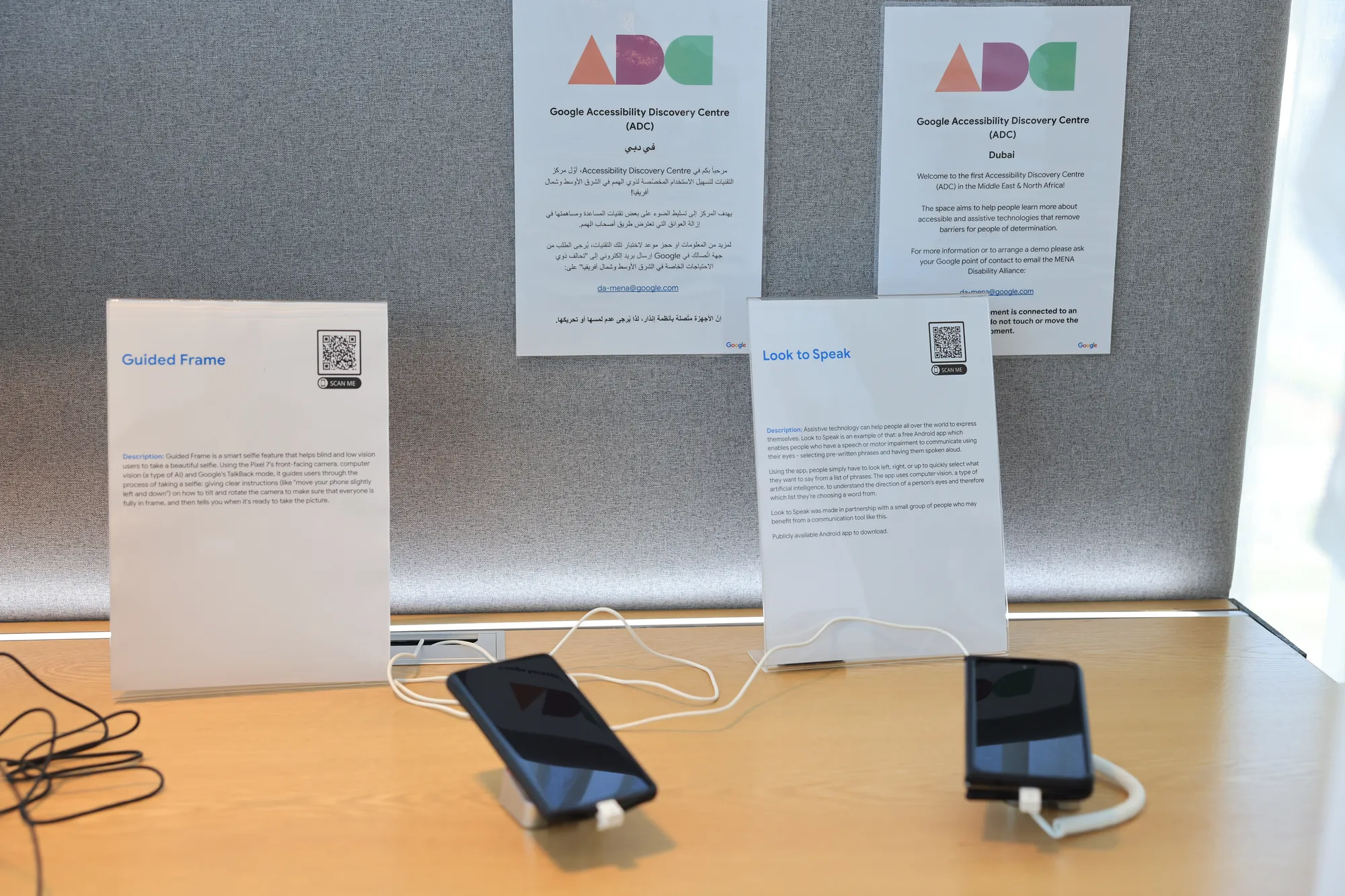 A table with two smartphones connected to stands. Each phone has informational posters behind it describing Google accessibility features "Guided Frame" and "Look to Speak." The posters include QR codes for more information.