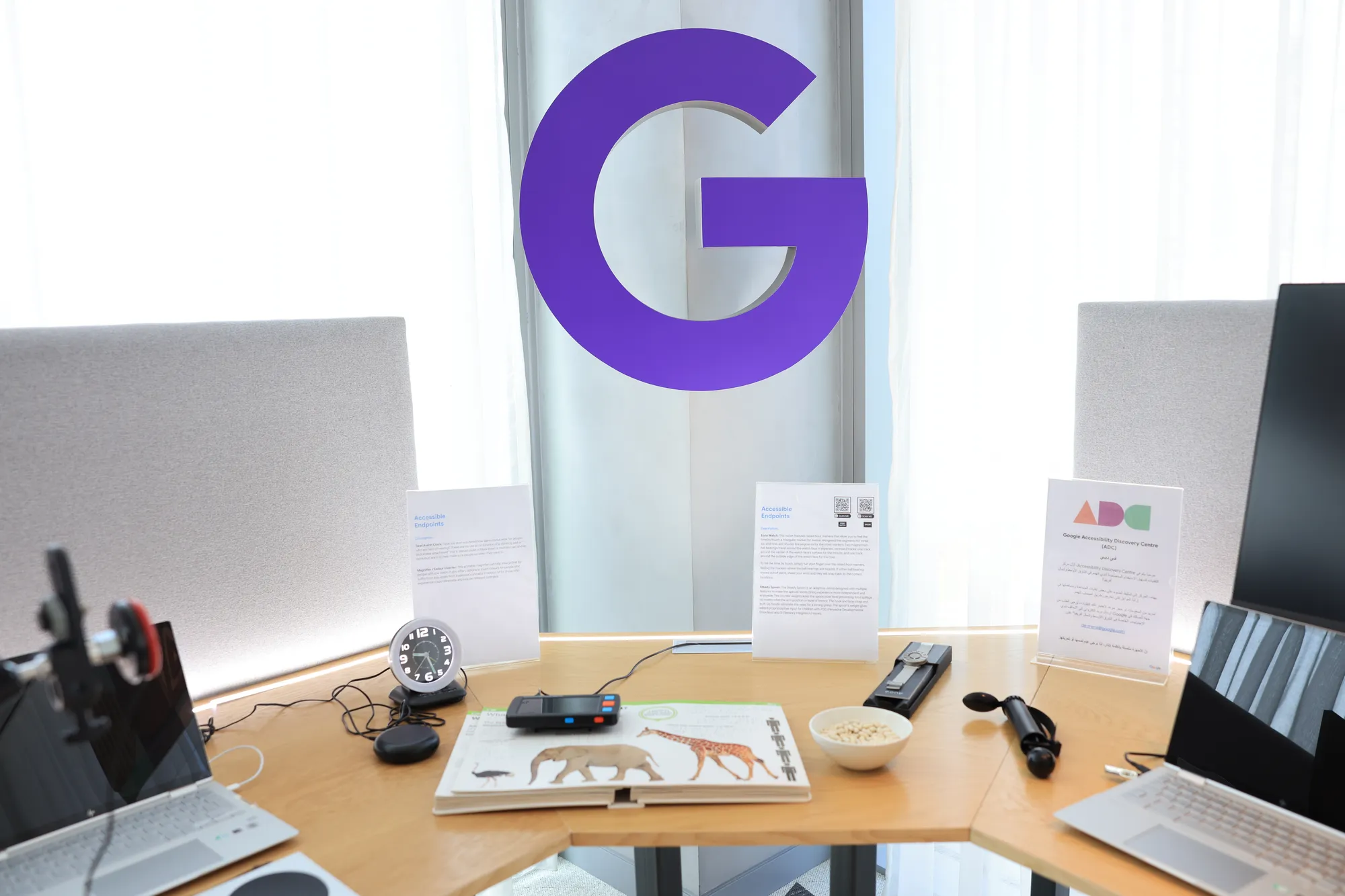 A wooden table with two laptops, a braille book for children, a deaf alarm clock and other electronic devices. A large purple letter "G" hangs in the background.