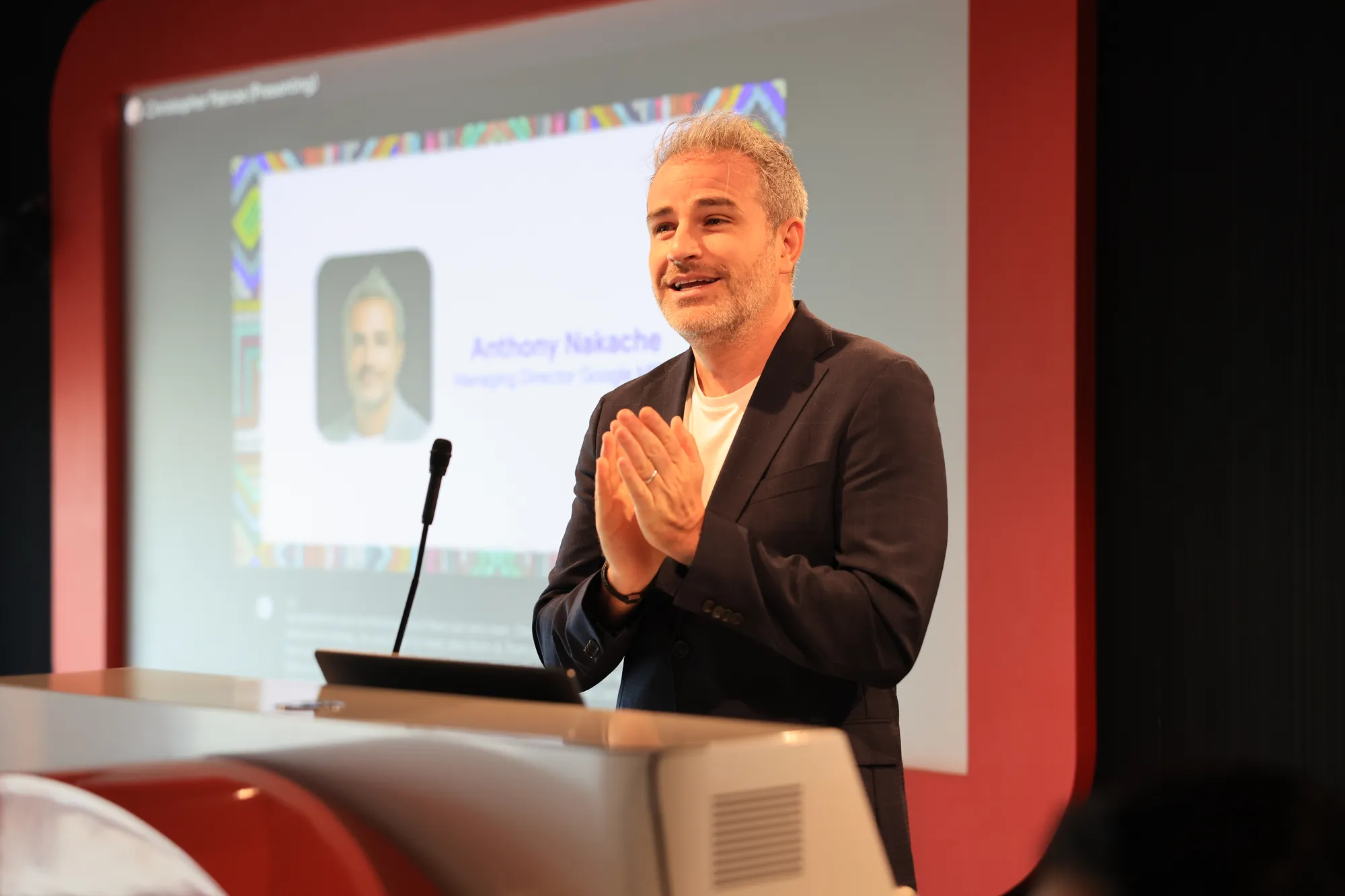 An image of a blonde male wearing a black suit jacket and white t-shirt speaking on a podium with a microphone in front of him. He has his hands together as he speaks. Behind him is a screen which has his name "Anthony Nakache" and his picture