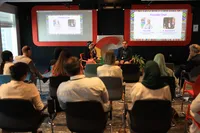 Image taken from the back of a room showing an audience and two speakers in a room with black walls and screens with red frames. The screens show the names of two people: Dalia Elfiki and Dr. Mostafa Attia. At the front, the two speakers are seated - one is a man and the other is a woman.