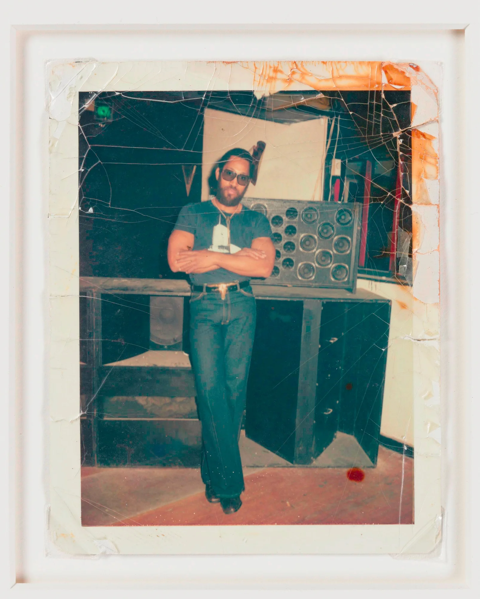 A color photograph of DJ Kool Herc in a white frame. In the photograph, which is slightly yellowed at the top right corner, DJ Kool Herc is standing in the middle of the frame with his arms crossed, wearing sunglasses with jeans and a t-shirt, looking directly at the viewer.