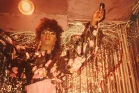 Drag queen Junior Labeija, in a black and pink floral blouse and colorful makeup, raises up her arms and a microphone while in front of a shimmery backdrop.