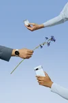 A photo of three hands against a blue background: One hand holds a Pixel 7a in Sea, another holds a blue flower and the third holds a pair of Pixel Buds A-Series in Sea.