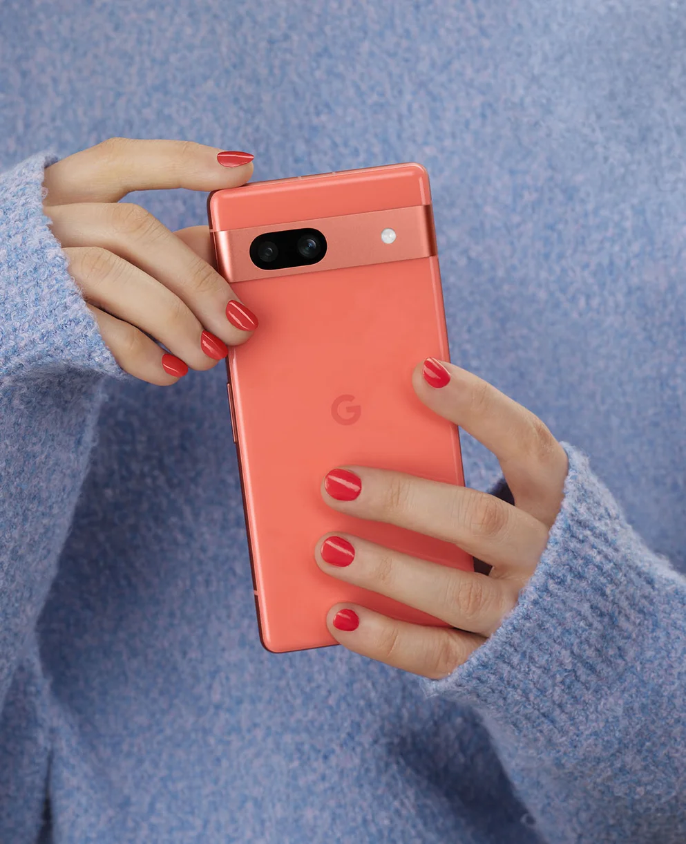 A photo showing a person’s hands holding a Pixel 7a. The person is wearing a blue sweater and their nails are painted coral, to match the coral Pixel 7a.