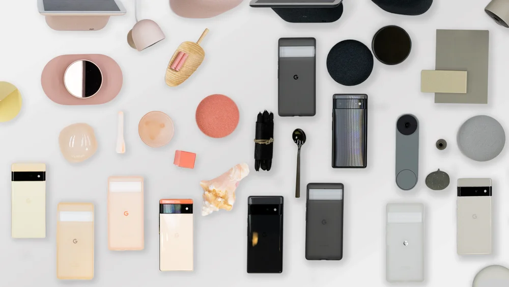 Overheard photograph of various items including Pixel 6 and 6 Pro phones in different shades of colors (mostly pink, white, yellow, and black hues) laid out on a white table in a gradient fashion.