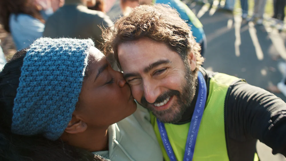 A bearded man wearing running gear and a medal smiles at the camera with a woman kissing his cheek