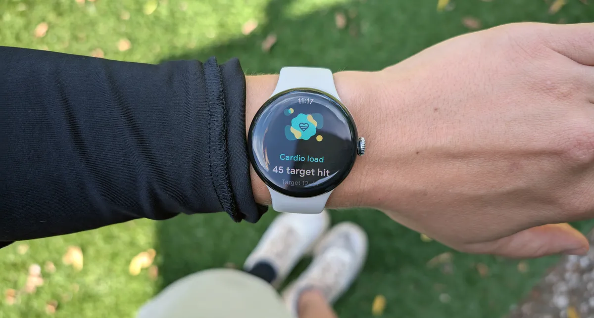 Photo showing a person's arm and wrist wearing a Pixel Watch 3. The display shows a heart icon and the words "Cardio Load. 45 target hit." In the background are slightly blurred tennis shoes and a green lawn.