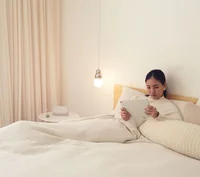 A photograph of a person sitting up in bed. They’re holding the Pixel tablet and there is a dock on the bedside table next to them.