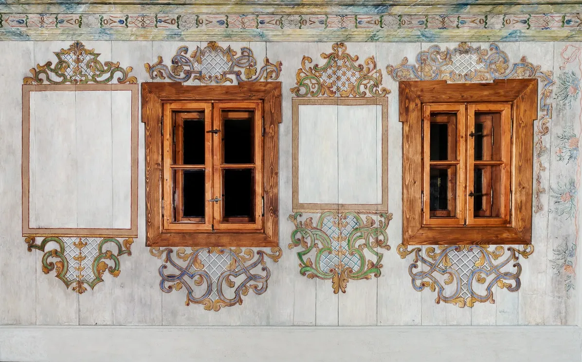 A light gray wall, in the middle of which there are two old wooden windows in a light brown, reddish color. Between the windows, there are two other windows painted on the wall, of the same size as the real ones. Ornaments in the form of swirls can be seen around the painted and real windows.