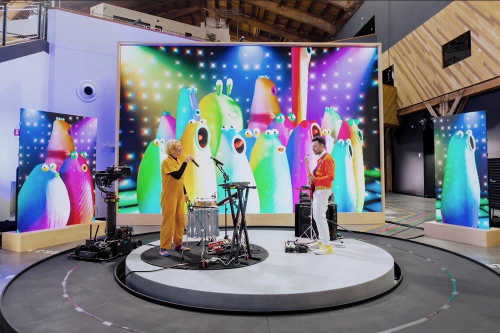 Two musicians play in front of a large screen, with the multi-colored animated “Blob Opera” singing on screen.