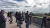 14 personas con sus teléfonos móviles toman fotos de un paisaje rural desde un mirador bajo un cielo nublado.