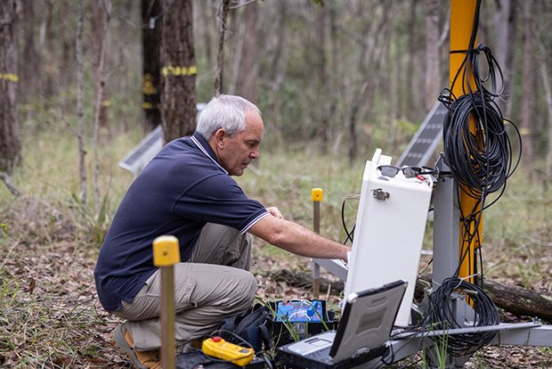 Professor Hamish McGowan, wearing a blue polo shirt and gray trousers, crouches in an area of green-brown woodland inspecting a laptop, radar and other equipment set up to detect bushfire risks.